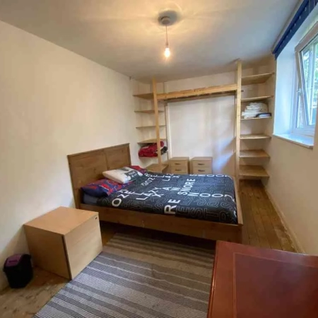 A spacious room with a double bed, bedding, window, storage, 2 drawers, a table, and a wall frame.