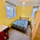 A room with a double bed, bedding, a table, a chair, 2 wardrobes, a light bulb, a yellow-coloured wall, and 2 windows.