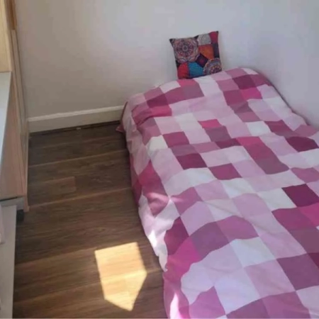 A room with a single bed featuring pink bedding, a wardrobe, and a table on the left side.