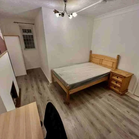 In this spacious Sydenham room, find a bed with a side table, a wall-mounted frame, a table and chair at the foot, and a window with a wardrobe at the other end.