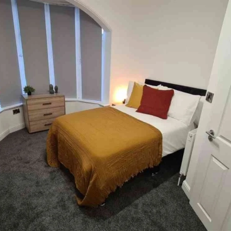 The bed is covered with white and yellow bedsheets, and pillows are placed on top. Beside the bed is a window and a small cabinet in this double room in Vauxhall, London.