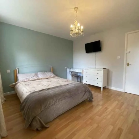 This furnished room in Vauxhall features a bed with bedding and pillows. On the right side, you'll find a table, chair, cabinet, and a wall-mounted TV, followed by a door.