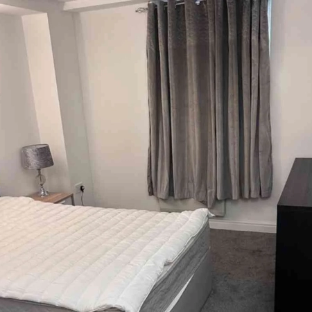This furnished room near Vauxhall Station features a bed, a bedside table with a lamp, a window adorned with a grey curtain, and another table.