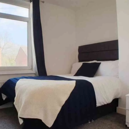 This double room near Streatham Station features a bed adorned with dark blue and white bedding, complemented by pillows on top. A window is situated on the left side, providing natural light and a pleasant atmosphere.