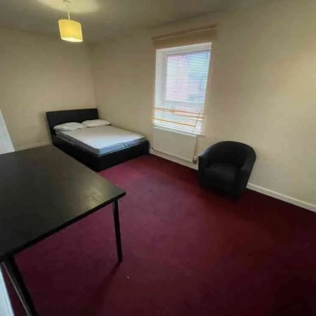 This furnished large double room features a fully red-carpeted floor. The bed is positioned in one corner, with a window beside it. Close to the bed, you'll find a single sofa for added comfort.