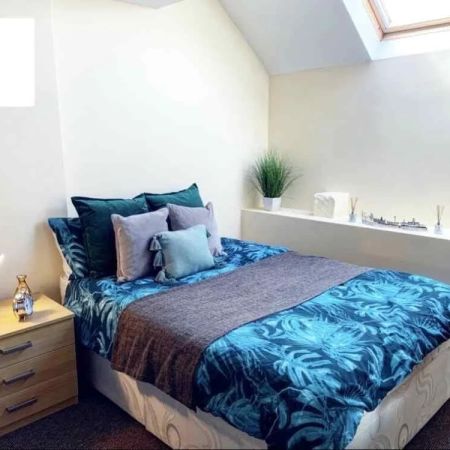 This double bedroom in Zone 2 has a bed with blue bedding, pillows and a side table with a lamp.