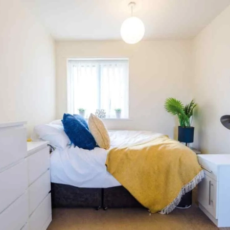 This furnished large room in Brixton features two cabinets, followed by a bed adorned with yellow bedding and pillows, positioned beside the window.