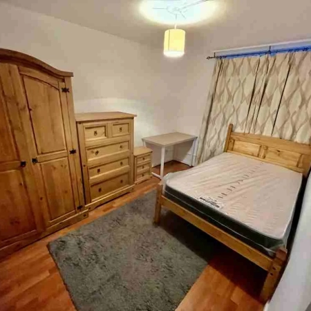 The stylish double room in Sydenham showcases a bed adorned with a curtain. Positioned on the left side is the table, followed by a side table, cabinet, and the wardrobe.