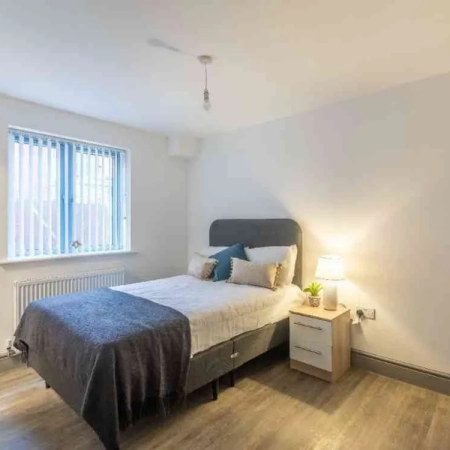 The furnished large room in Vauxhall features a bed adorned with bedding and topped with pillows. A window is situated on the left side, and a side table with a lamp is placed on the right.