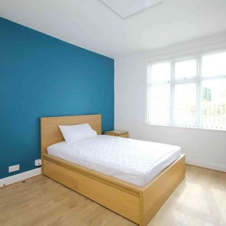 This fully furnished spacious room for rent features a blue wall behind the bed adorned with bedding, and a side table on the right, leading to the window.