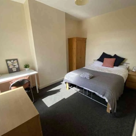 This central London room for rent features a bed with bedding and pillows. To the right, you'll find a sidetable, and on the left, there's a wardrobe. Opposite the bed is a window, a table, a chair, and a cabinet.