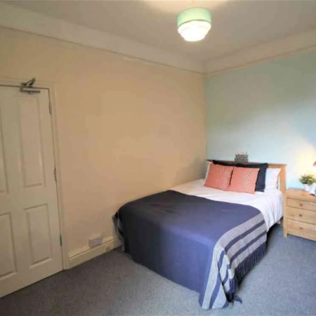 This Battersea furnished room for rent features a bed adorned with white and blue bedding and pillows. A side table is positioned on the right, and the door is located at the foot of the bed.