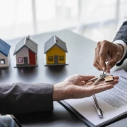 A hand passing a key to another hand against the backdrop of three display houses in the 'Landlord's Success with Rent-Guaranteed' blog