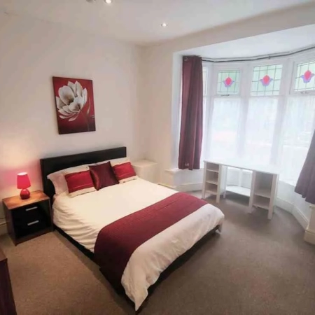A room with a double bed, bedding, pillows, curtained large windows, a bedside table with a lamp and cabinets.