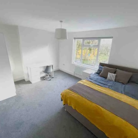 This spacious room near Tower Bridge features a bed with bedding and pillows, accompanied by a side table on the left, followed by a window. Adjacent to the window is the table and chair, and further along is the white wardrobe.